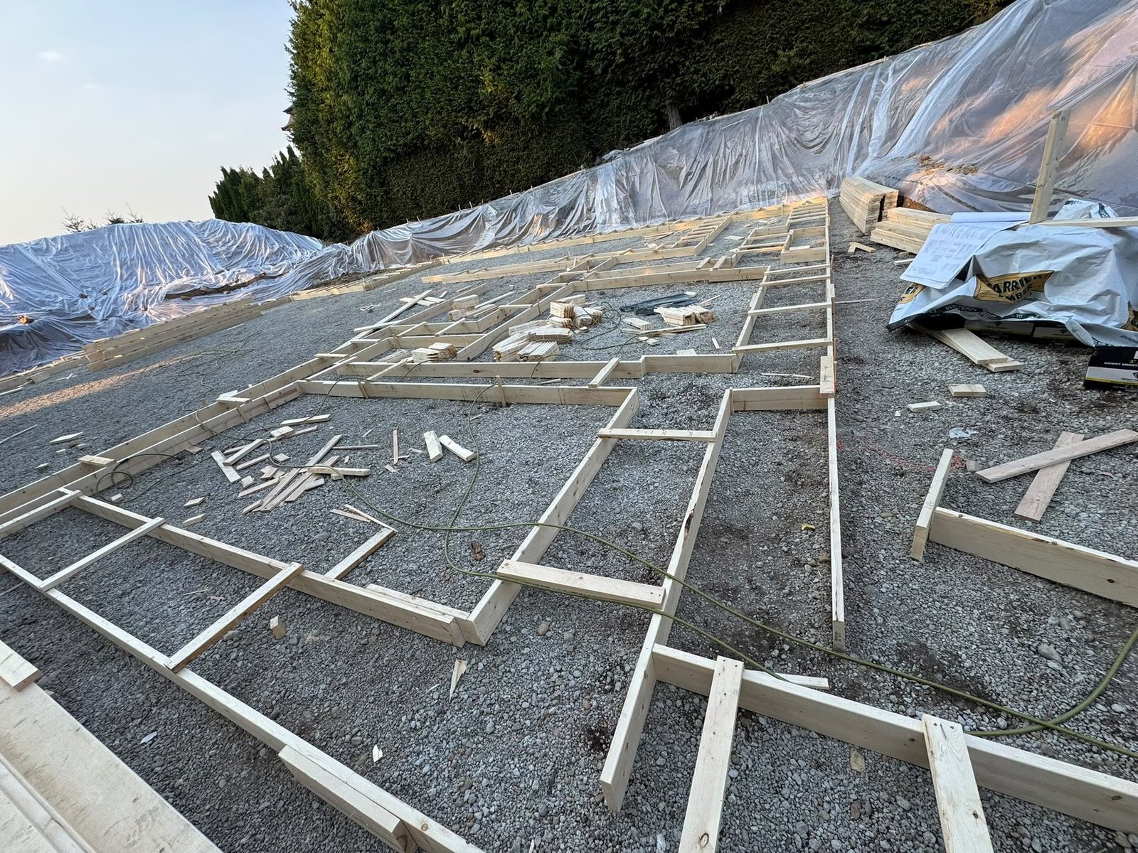 Wood layout forms across a gravel construction area, with lumber stacks under protective sheeting.