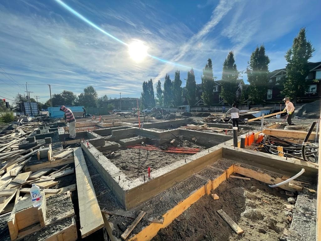 Workers beside formed concrete foundation walls and exposed service pipes on a residential site.
