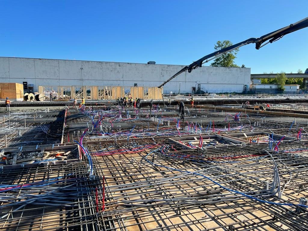 Concrete pump boom over a reinforced slab with coloured service lines and wood-framed walls beyond.