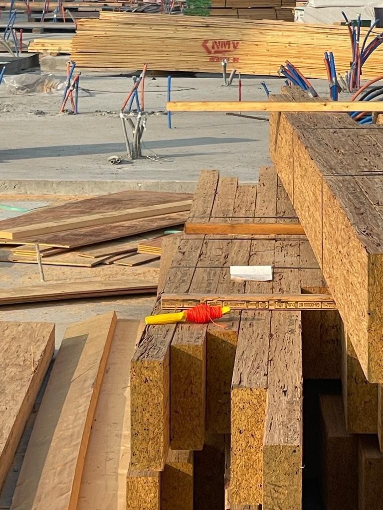Stacked engineered wood members and dimensional lumber beside a concrete work area with service pipes.