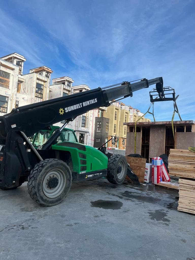 Telehandler lifting a worker and materials beside an exterior wood-framed wall.
