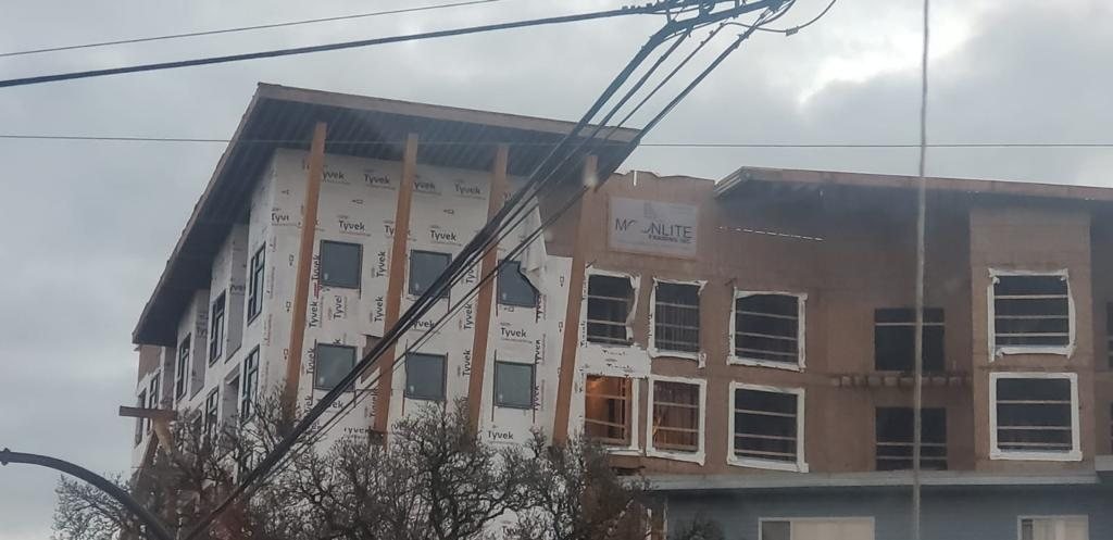 Wood-framed multi-unit building viewed through roadside trees and utility lines.
