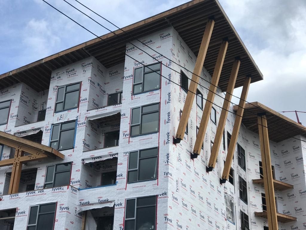 Exposed timber canopy supports beside a multi-storey wall covered in weather protection.